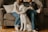 Two people are sitting on a couch, one holding a cat, while a large white dog is sitting in front of them. The setting appears cozy with pillows and a wooden floor visible. The mood suggests a relaxed, domestic atmosphere.