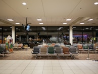 An airport waiting area with rows of empty seats in various colors, large windows showing a nighttime view of city lights, and a television suspended from the ceiling. A children's play area is visible on the left with interactive games.