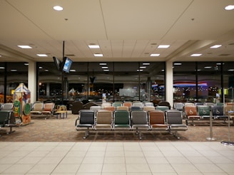 An airport waiting area with rows of empty seats in various colors, large windows showing a nighttime view of city lights, and a television suspended from the ceiling. A children's play area is visible on the left with interactive games.