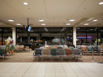 An airport waiting area with rows of empty seats in various colors, large windows showing a nighttime view of city lights, and a television suspended from the ceiling. A children's play area is visible on the left with interactive games.