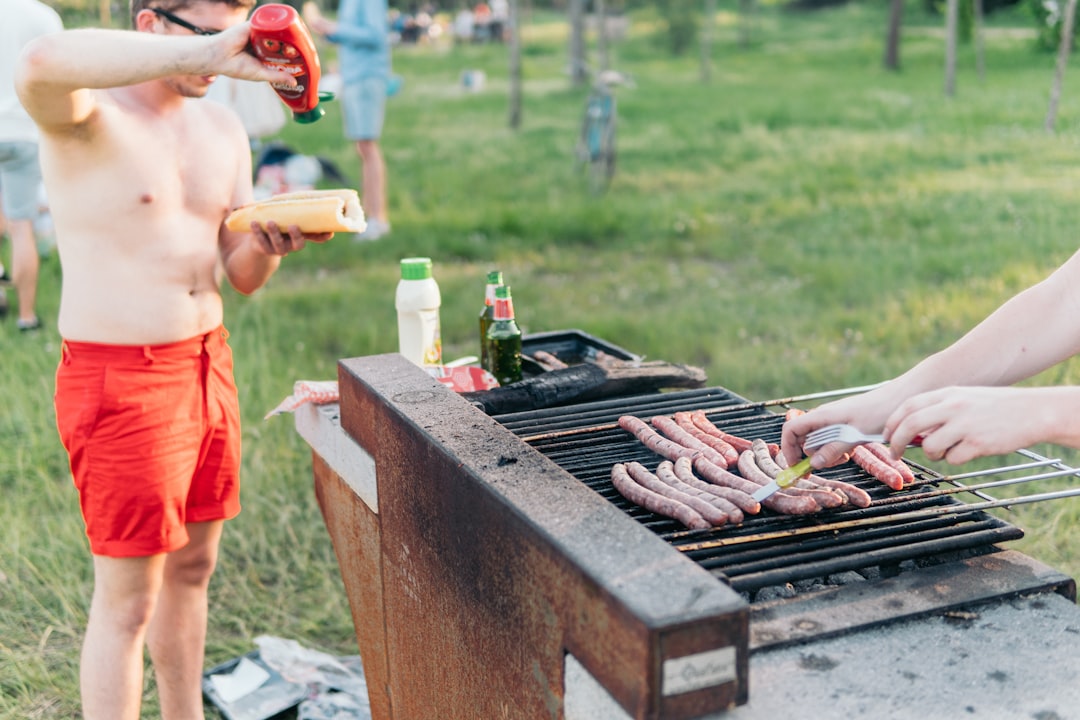 man wearing red shorts standing at grill putting ketchup on hotdog