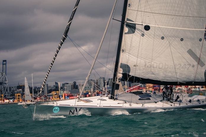 A large sailboat with a sleek hull navigates through choppy waters in a busy harbor setting. The background is lined with industrial cargo areas and cranes, and the city skyline is visible under a cloudy sky. The sailboat is equipped with large sails and features multiple crew members handling the vessel.
