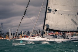 A large sailboat with a sleek hull navigates through choppy waters in a busy harbor setting. The background is lined with industrial cargo areas and cranes, and the city skyline is visible under a cloudy sky. The sailboat is equipped with large sails and features multiple crew members handling the vessel.