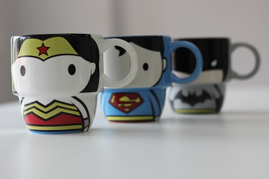 Three ceramic mugs with cartoon-style designs, each featuring a different superhero emblem and color theme, are arranged in a row on a white surface. The mugs represent popular characters with distinctive traits, including a red and yellow star emblem, an 'S' symbol on a blue background, and a black bat symbol on gray.