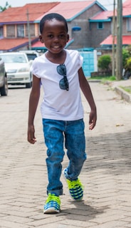 A young child is walking confidently on a paved street. The child is smiling, wearing a white T-shirt with sunglasses clipped to it, blue jeans, and brightly colored sneakers. In the background, there are houses with red roofs, and a row of parked cars lines the street.