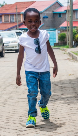 A young child is walking confidently on a paved street. The child is smiling, wearing a white T-shirt with sunglasses clipped to it, blue jeans, and brightly colored sneakers. In the background, there are houses with red roofs, and a row of parked cars lines the street.