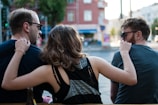 Three people are sitting together outdoors, with their backs towards the camera. They appear to be engaging in a relaxed, friendly conversation. The person in the middle is playfully putting an arm around the other two. They are wearing casual clothing and sunglasses, and an urban environment with blurry buildings in the background suggests a city setting.