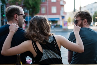 Three people are sitting together outdoors, with their backs towards the camera. They appear to be engaging in a relaxed, friendly conversation. The person in the middle is playfully putting an arm around the other two. They are wearing casual clothing and sunglasses, and an urban environment with blurry buildings in the background suggests a city setting.