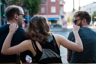 Three people are sitting together outdoors, with their backs towards the camera. They appear to be engaging in a relaxed, friendly conversation. The person in the middle is playfully putting an arm around the other two. They are wearing casual clothing and sunglasses, and an urban environment with blurry buildings in the background suggests a city setting.