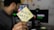 A person is holding a yellow sticky note with the word 'BLOCKCHAIN' written on it. In the background, a computer monitor displays a blurry image, and there is an energetic focus on the note.