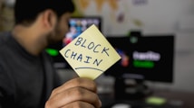 A person is holding a yellow sticky note with the word 'BLOCKCHAIN' written on it. In the background, a computer monitor displays a blurry image, and there is an energetic focus on the note.