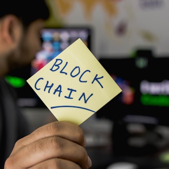 A person is holding a yellow sticky note with the word 'BLOCKCHAIN' written on it. In the background, a computer monitor displays a blurry image, and there is an energetic focus on the note.