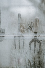 A foggy or frosted glass surface with outlines of gardening tools visible through it. The tools seem to be a shovel, rake, and hoe, with handles upright. The frosted glass creates a blurred and muted view of these objects, adding a mysterious and ethereal quality.