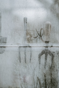 A foggy or frosted glass surface with outlines of gardening tools visible through it. The tools seem to be a shovel, rake, and hoe, with handles upright. The frosted glass creates a blurred and muted view of these objects, adding a mysterious and ethereal quality.