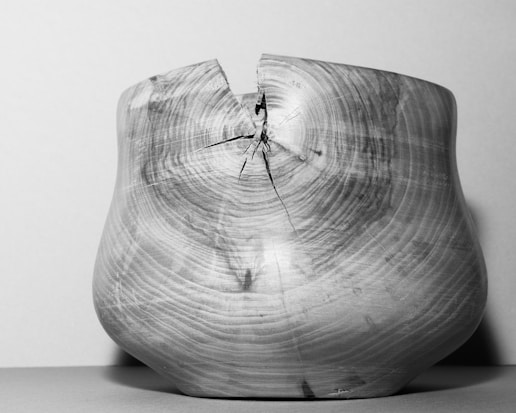 A wooden bowl with a prominent crack at the top, showcasing natural wood grain patterns. The bowl's simple, rounded shape contrasts with its asymmetric defect, highlighting its rustic and handmade qualities.