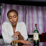 A woman with short hair and vibrant makeup sits at a table, wearing a white lace-trimmed robe. She has her fingers gently resting on a glass of whiskey. A bottle of Jameson Irish Whiskey is prominently placed next to her on the table. The background features pink and white curtains, casting a soft ambiance.