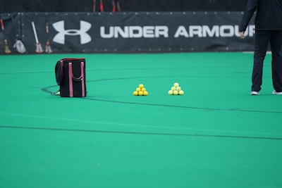 A sports field features a black and pink sports bag, two pyramids of yellow balls, and a portion of a person in dark athletic gear. The background displays a banner with the Under Armour logo.