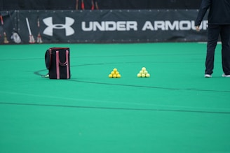 A sports field features a black and pink sports bag, two pyramids of yellow balls, and a portion of a person in dark athletic gear. The background displays a banner with the Under Armour logo.