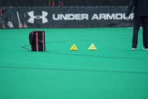 A sports field features a black and pink sports bag, two pyramids of yellow balls, and a portion of a person in dark athletic gear. The background displays a banner with the Under Armour logo.