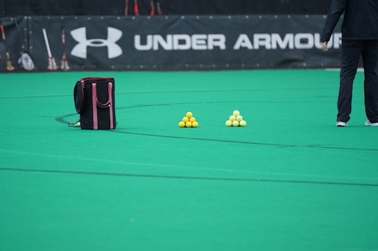 A sports field features a black and pink sports bag, two pyramids of yellow balls, and a portion of a person in dark athletic gear. The background displays a banner with the Under Armour logo.