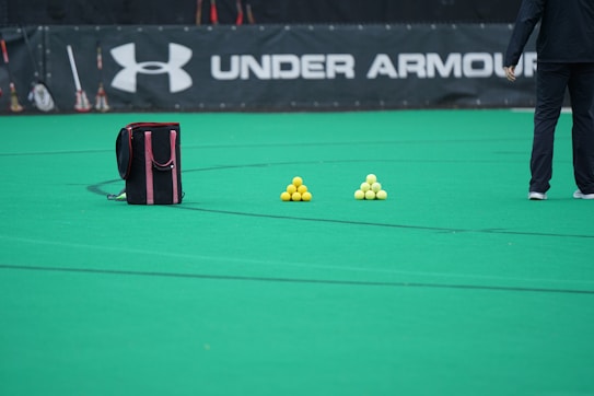 A sports field features a black and pink sports bag, two pyramids of yellow balls, and a portion of a person in dark athletic gear. The background displays a banner with the Under Armour logo.