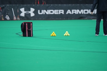 A sports field features a black and pink sports bag, two pyramids of yellow balls, and a portion of a person in dark athletic gear. The background displays a banner with the Under Armour logo.