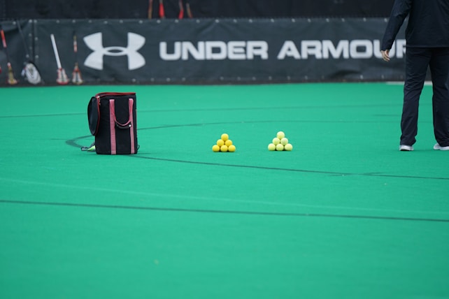 A sports field features a black and pink sports bag, two pyramids of yellow balls, and a portion of a person in dark athletic gear. The background displays a banner with the Under Armour logo.