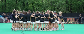 A group of female athletes in black and white sports uniforms huddle together on a green field, surrounded by lush green trees in the background. They appear to be having a team meeting or pep talk before a game.