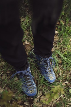 Blue hiking boots with thick laces are stepping on grass and dried leaves. The scene captures a woodland or nature setting with various shades of green foliage.