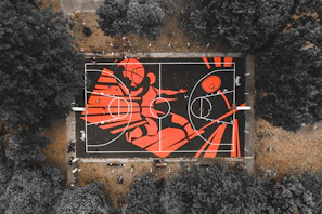 An aerial view of a basketball court with a striking mural featuring bold red and black colors. The mural depicts a stylized figure in an action pose. Surrounding the court are trees with muted colors creating a dramatic contrast. People can be seen on the court, playing or observing.
