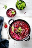 A pot of vibrant red borscht, garnished with fresh herbs, is accompanied by a smaller bowl of the same soup and a bowl of green leafy vegetables. A spoon with a small amount of borscht lies next to a dark napkin.