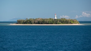 A small island with dense vegetation is surrounded by blue ocean waters. A white lighthouse with a red top stands prominently on the island, overlooking the sea. The sky is partly cloudy, and there are distant mountains visible on the horizon.