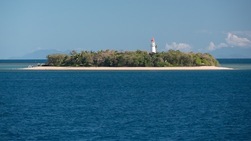 A small island with dense vegetation is surrounded by blue ocean waters. A white lighthouse with a red top stands prominently on the island, overlooking the sea. The sky is partly cloudy, and there are distant mountains visible on the horizon.