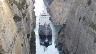 A large cargo ship is navigating a narrow canal with steep rocky walls rising on either side. The canal is a tight fit for the vessel, emphasizing the immense size of the ship in contrast to the narrow passage.
