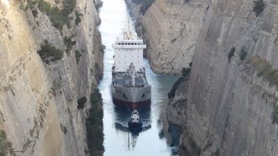 A large cargo ship is navigating a narrow canal with steep rocky walls rising on either side. The canal is a tight fit for the vessel, emphasizing the immense size of the ship in contrast to the narrow passage.