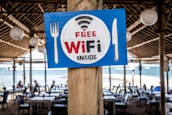 A wooden sign featuring the words 'Free WiFi Inside' is prominently displayed on a wooden pillar. The sign has a blue background with a white circle, and the text is in red and black. It is decorated with symbols of a fork on the left and a knife on the right. In the background, there is an outdoor dining area with tables and chairs under a thatched roof, and a view of the sea is visible.