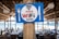 A wooden sign featuring the words 'Free WiFi Inside' is prominently displayed on a wooden pillar. The sign has a blue background with a white circle, and the text is in red and black. It is decorated with symbols of a fork on the left and a knife on the right. In the background, there is an outdoor dining area with tables and chairs under a thatched roof, and a view of the sea is visible.