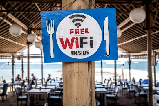 A wooden sign featuring the words 'Free WiFi Inside' is prominently displayed on a wooden pillar. The sign has a blue background with a white circle, and the text is in red and black. It is decorated with symbols of a fork on the left and a knife on the right. In the background, there is an outdoor dining area with tables and chairs under a thatched roof, and a view of the sea is visible.