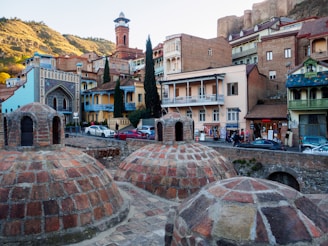 A scenic view of a historic area featuring old brick structures with domed rooftops in the foreground. These domes are part of what appears to be an ancient bathhouse. In the background, there are various colorful buildings with balconies, ornate detailing, and medieval architecture. A hill with greenery and ancient fortress walls is visible in the distance, enhancing the historical atmosphere of the scene.