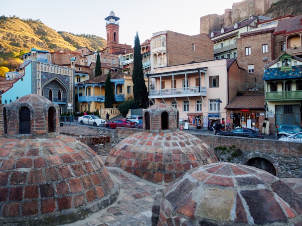 A scenic view of a historic area featuring old brick structures with domed rooftops in the foreground. These domes are part of what appears to be an ancient bathhouse. In the background, there are various colorful buildings with balconies, ornate detailing, and medieval architecture. A hill with greenery and ancient fortress walls is visible in the distance, enhancing the historical atmosphere of the scene.