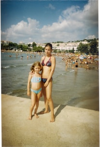 Two people wearing swimsuits stand on a concrete surface near a beach. The background is filled with numerous individuals enjoying a day by the sea, with some swimming in the water and others lounging on the sand. Buildings and trees are visible in the distance under a partly cloudy sky.