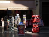 A storefront displaying traditional Spanish dresses in red and black with lace elements. Several racks hold postcards, accessories, and souvenirs, with patterned dresses hanging prominently. The store roof says 'Mantones y Mantillas' and a passerby is partially visible.
