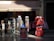 A storefront displaying traditional Spanish dresses in red and black with lace elements. Several racks hold postcards, accessories, and souvenirs, with patterned dresses hanging prominently. The store roof says 'Mantones y Mantillas' and a passerby is partially visible.