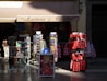 A storefront displaying traditional Spanish dresses in red and black with lace elements. Several racks hold postcards, accessories, and souvenirs, with patterned dresses hanging prominently. The store roof says 'Mantones y Mantillas' and a passerby is partially visible.