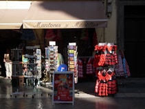 A storefront displaying traditional Spanish dresses in red and black with lace elements. Several racks hold postcards, accessories, and souvenirs, with patterned dresses hanging prominently. The store roof says 'Mantones y Mantillas' and a passerby is partially visible.
