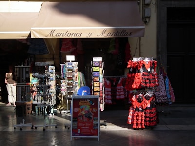 A storefront displaying traditional Spanish dresses in red and black with lace elements. Several racks hold postcards, accessories, and souvenirs, with patterned dresses hanging prominently. The store roof says 'Mantones y Mantillas' and a passerby is partially visible.