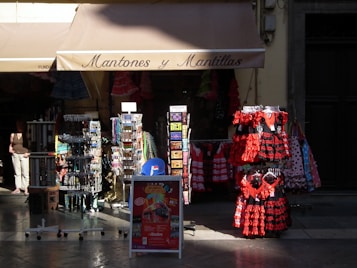 A storefront displaying traditional Spanish dresses in red and black with lace elements. Several racks hold postcards, accessories, and souvenirs, with patterned dresses hanging prominently. The store roof says 'Mantones y Mantillas' and a passerby is partially visible.