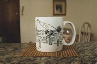 A white mug with a black line drawing depicting a cityscape, featuring buildings, people, and palm trees, is placed on a wooden mat on a granite countertop. In the blurred background, part of a chair, a laptop, and a framed picture on the wall are visible.