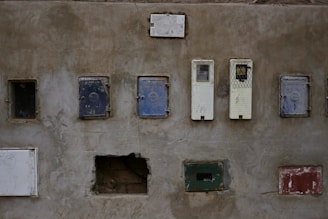 A weathered concrete wall featuring multiple electrical panels or meter boxes arranged in two horizontal rows. The panels vary in color and condition, with some appearing rusty or worn. The wall itself shows signs of age with cracks and peeling surfaces.