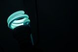 A glowing compact fluorescent light bulb with a spiral shape stands out against a dark background, emitting a soft teal light.